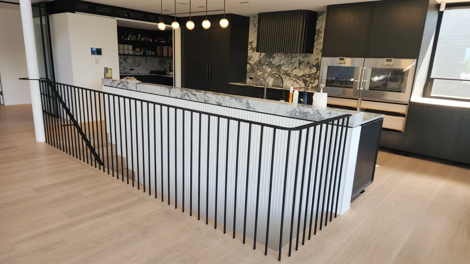 Black steel vertical-bar balustrade around a stair void beside a marble kitchen.