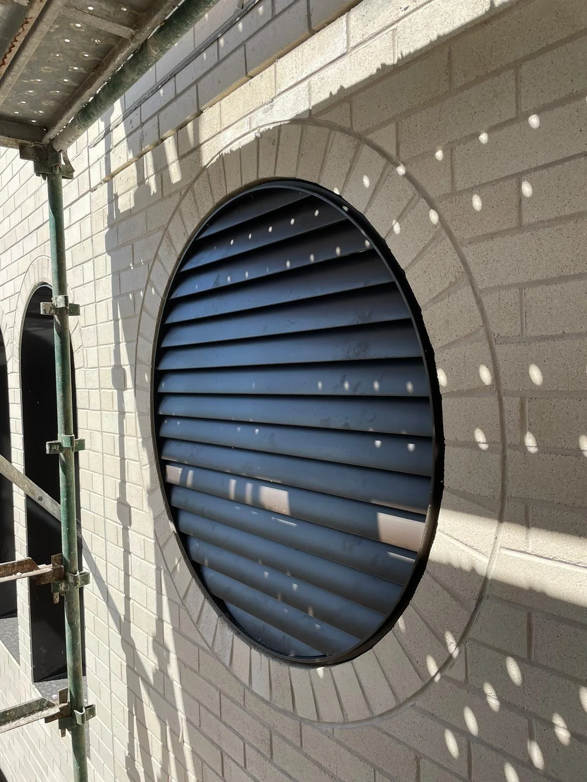Circular louvred steel window screen set into a brick wall.