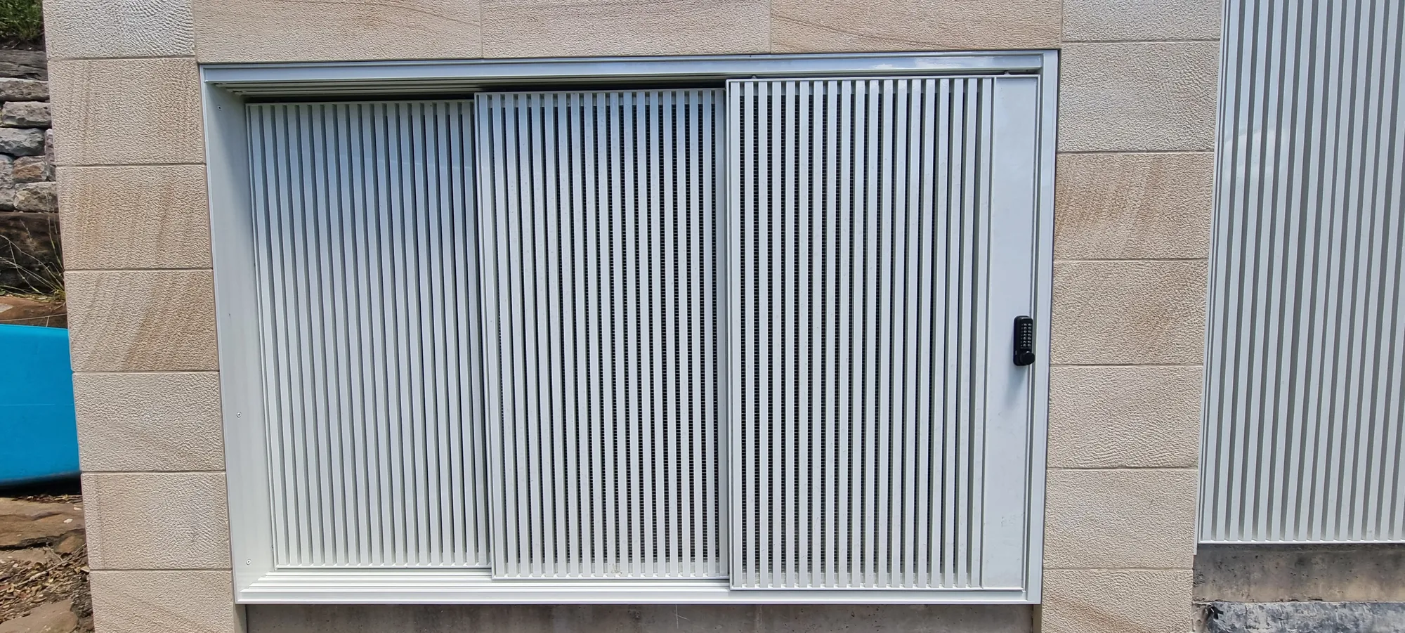 Light-grey aluminium vertical-batten sliding gate on a sandstone wall.