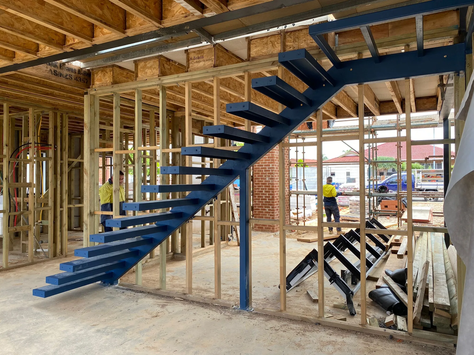 Blue mono-stringer steel staircase with folded-plate treads being installed in a timber-framed new build.