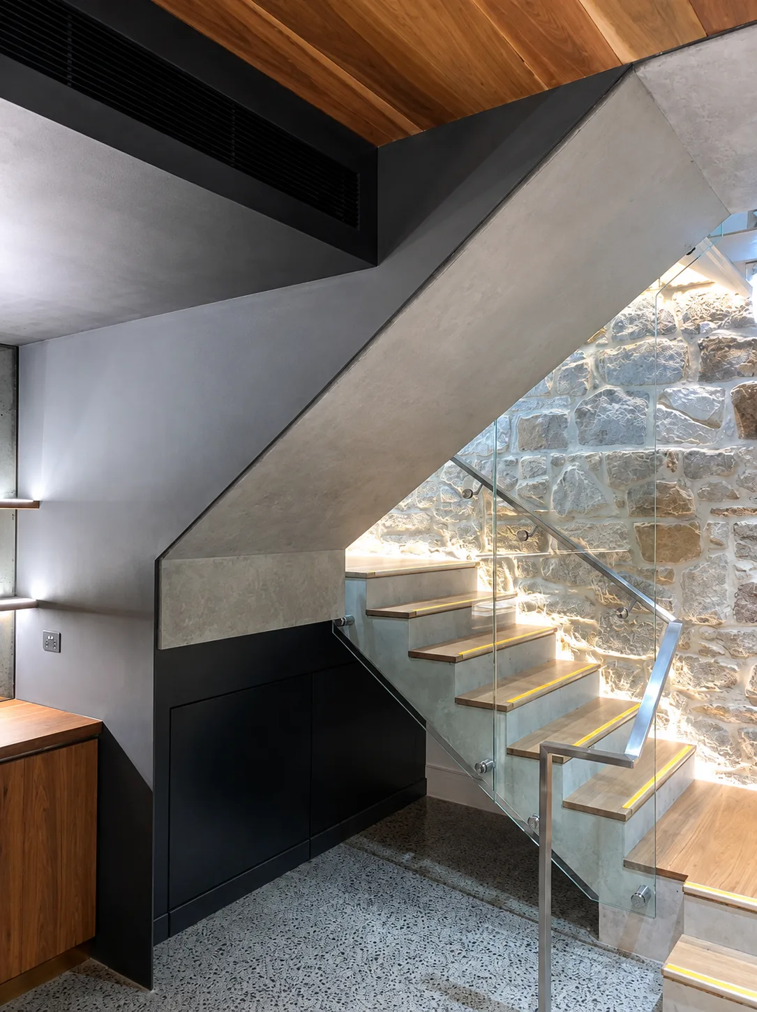 Folded-plate steel staircase with glass balustrade against a sandstone wall.