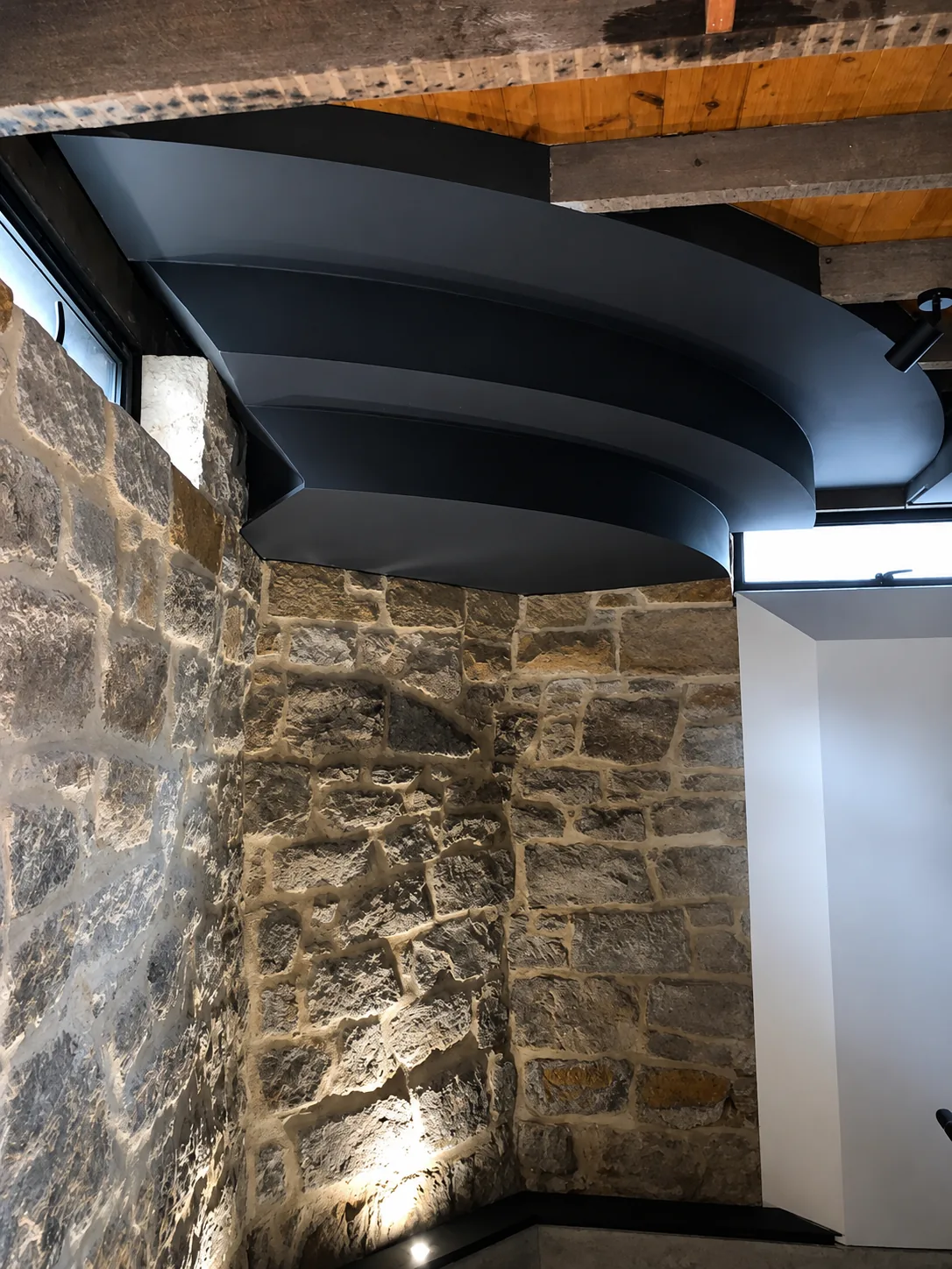 Underside detail of a curved blackened-steel balcony over a sandstone wall.