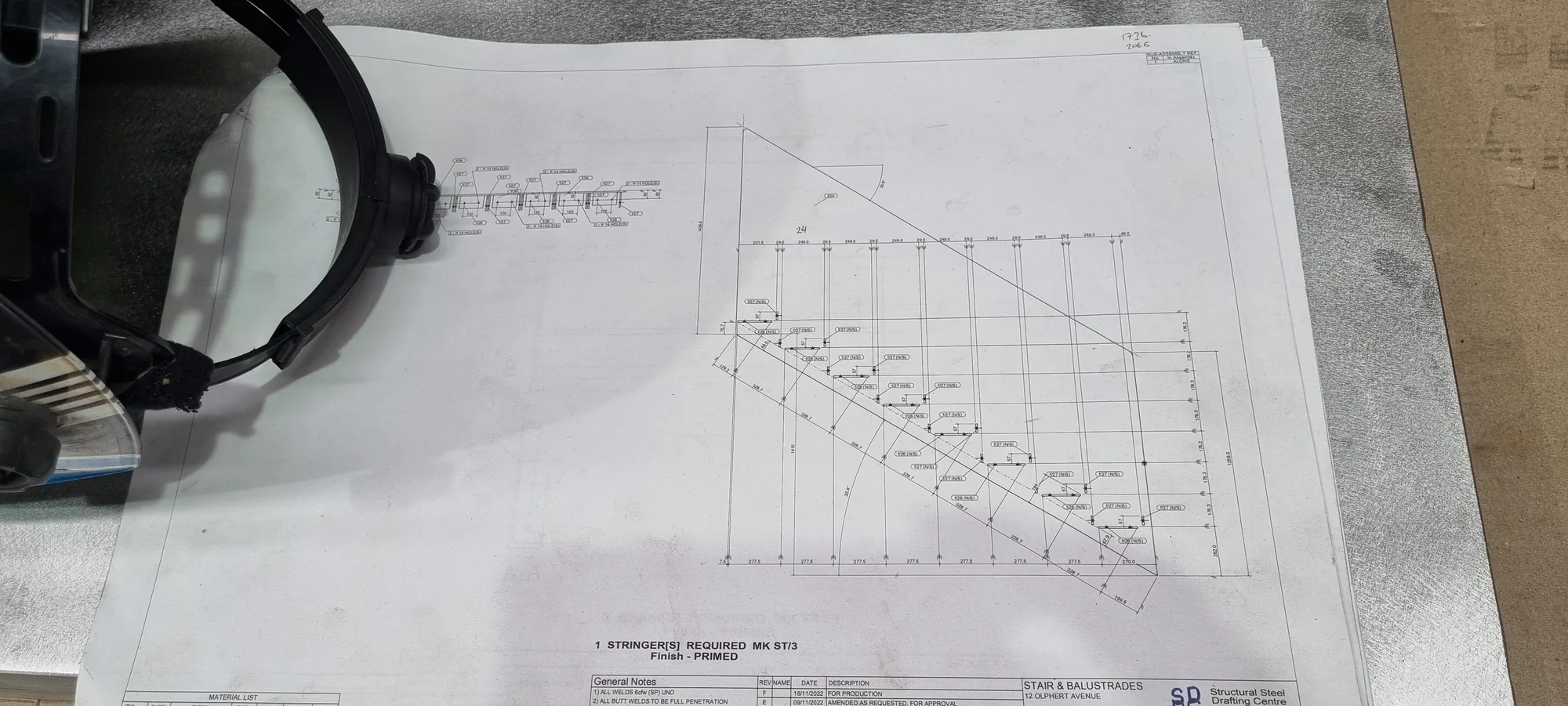 A staircase shop drawing and engineering plan on the bench with a welding mask.