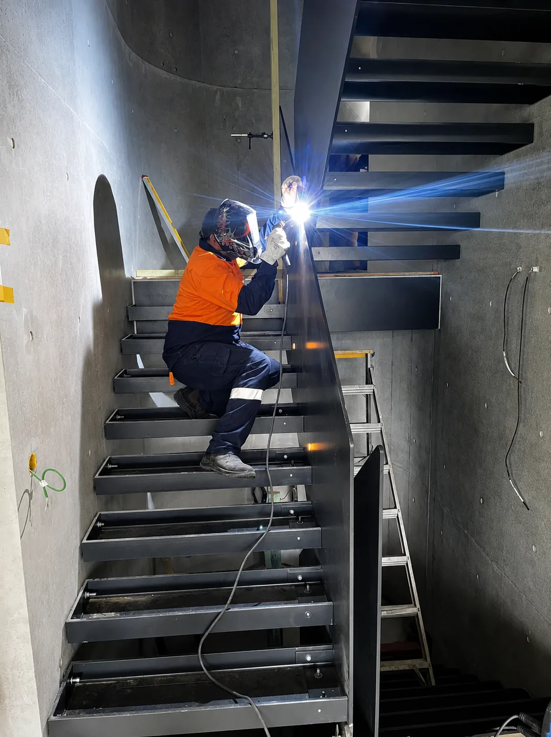 A welder in full PPE welding a steel staircase on site, the arc lighting the scene.